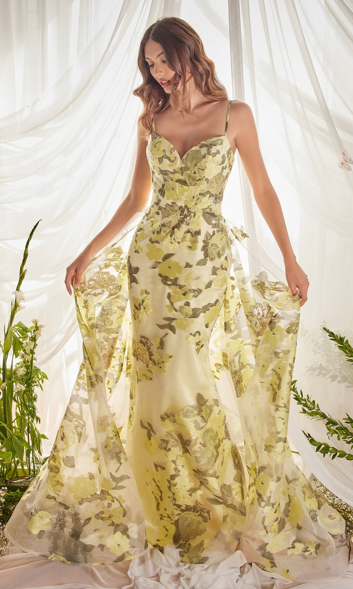 A woman wearing the Lemon Yellow Floral-Print Long Prom Dress A0770, a fitted strapless gown with yellow and green floral accents, stands before sheer white curtains and holds out the flowing sheer fabric of her dress.